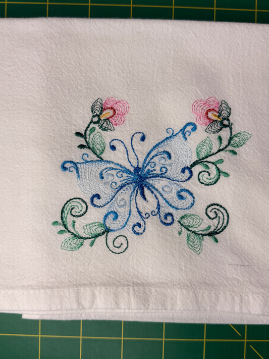 A white fabric kitchen towel featuring an embroidered design of a blue butterfly with pink flowers and green leaves, laid flat on a green cutting mat.