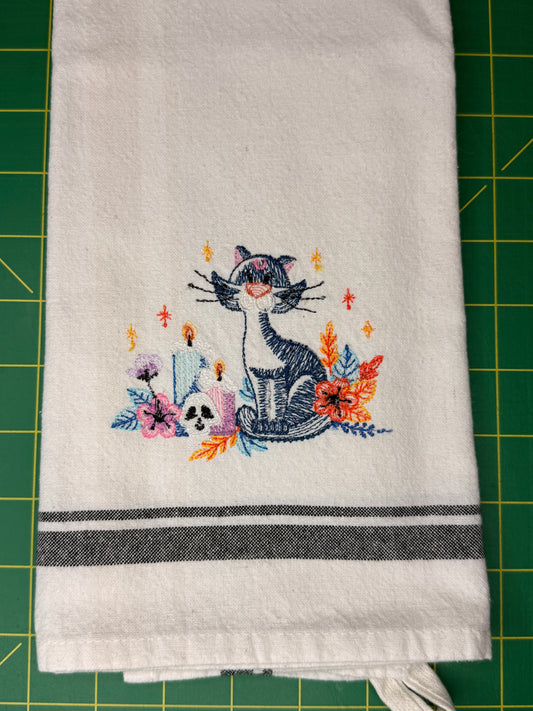A white kitchen towel with a colorful embroidered design of a cat, flowers, and candles, featuring two dark gray horizontal stripes near the bottom, laid flat on a green cutting mat.