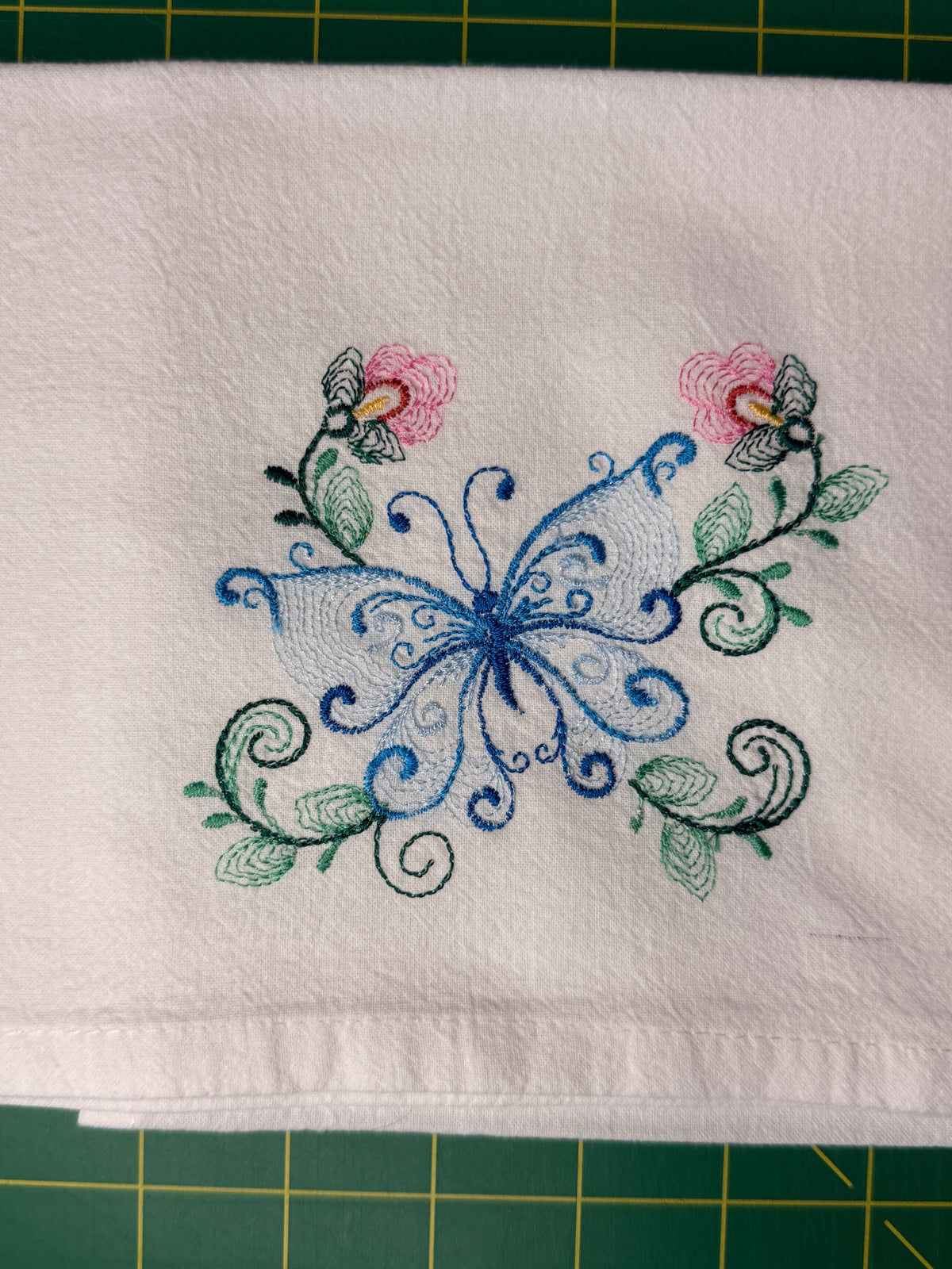 A white fabric kitchen towel featuring an embroidered design of a blue butterfly with pink flowers and green leaves, laid flat on a green cutting mat.