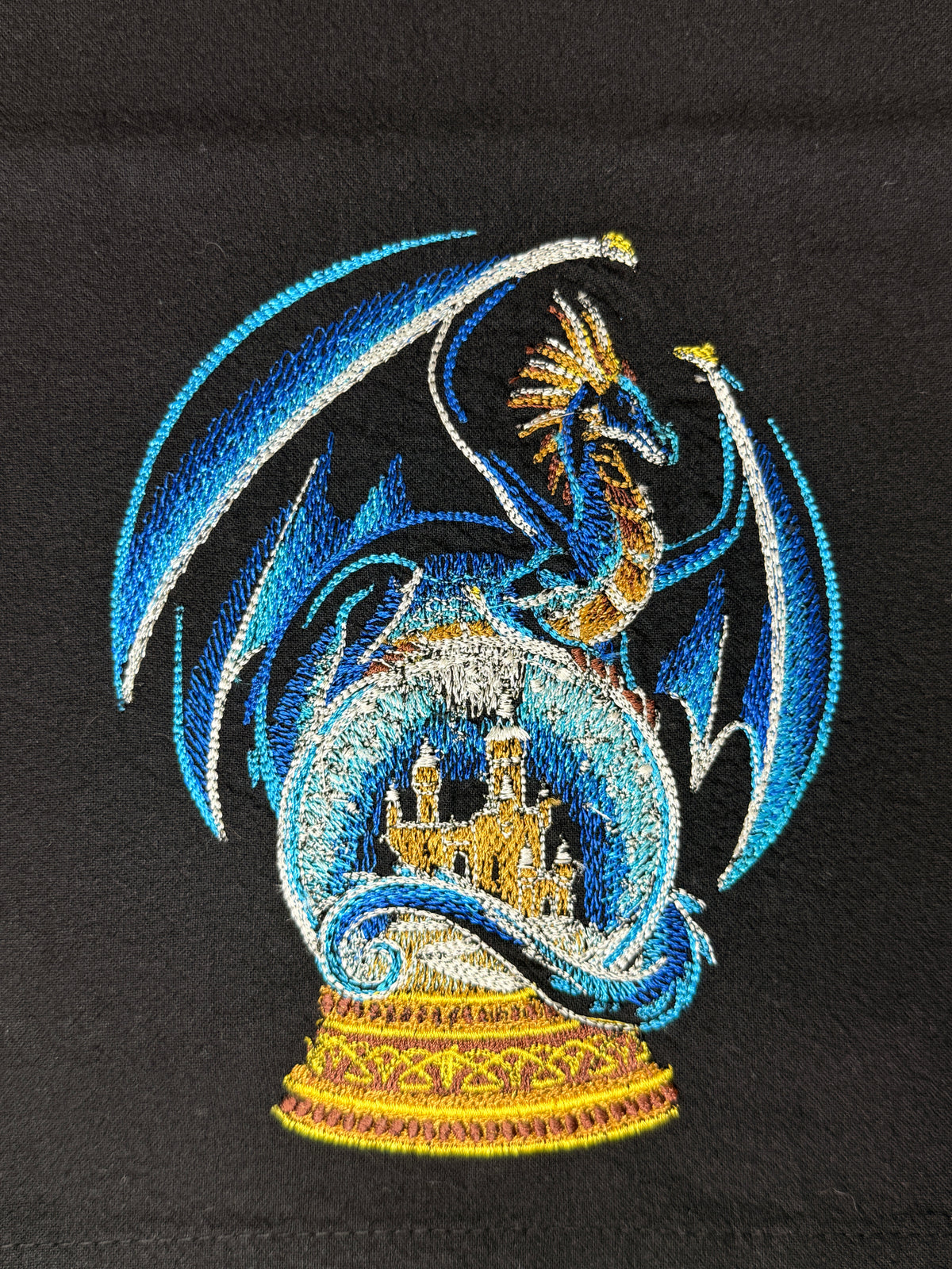 A close-up of a detailed embroidered design featuring a blue dragon with gold accents surrounding a castle inside a circular frame, set against a black fabric background.
