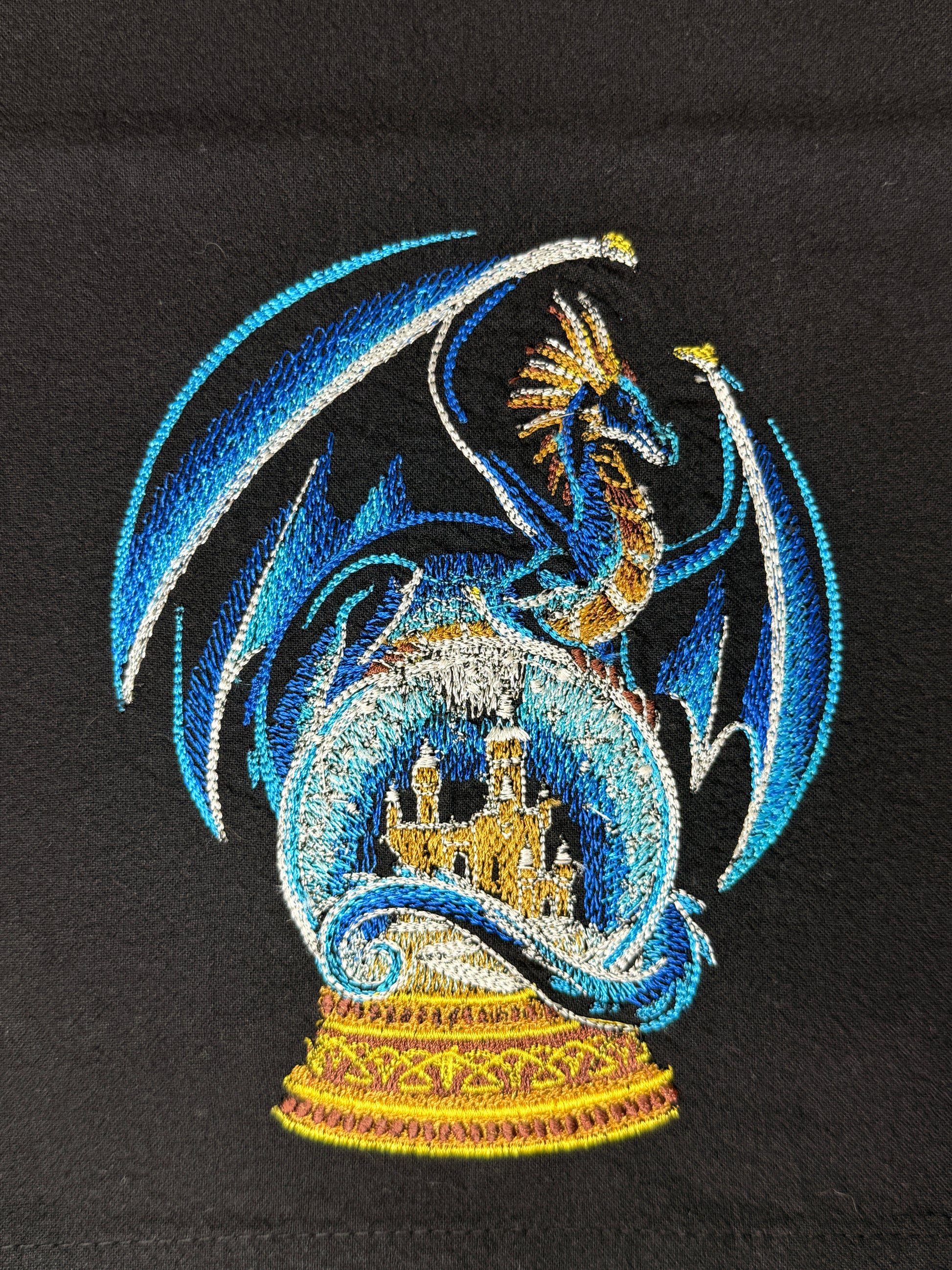 A close-up of a detailed embroidered design featuring a blue dragon with gold accents surrounding a castle inside a circular frame, set against a black fabric background.