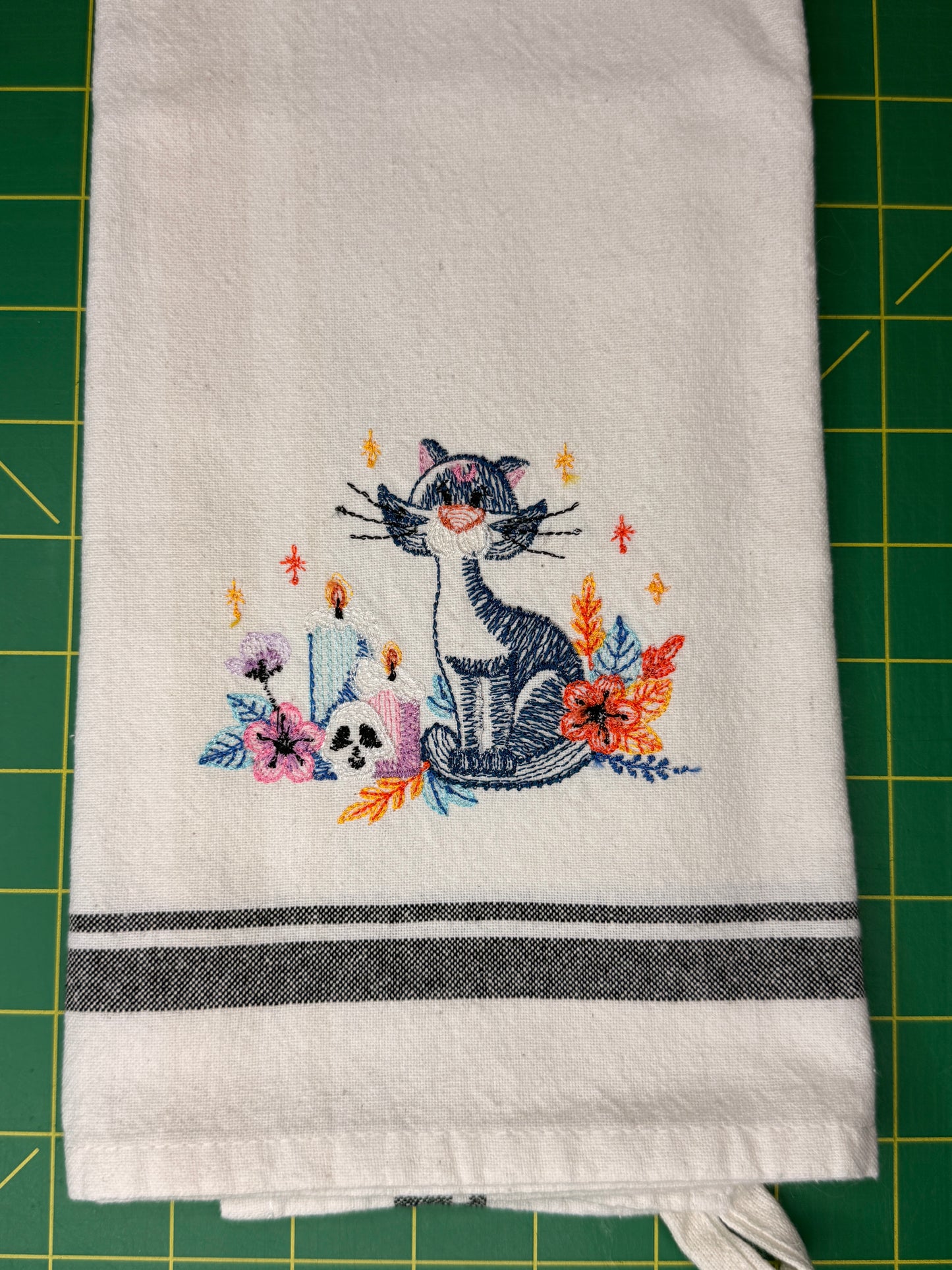 A white kitchen towel with a colorful embroidered design of a cat, flowers, and candles, featuring two dark gray horizontal stripes near the bottom, laid flat on a green cutting mat.