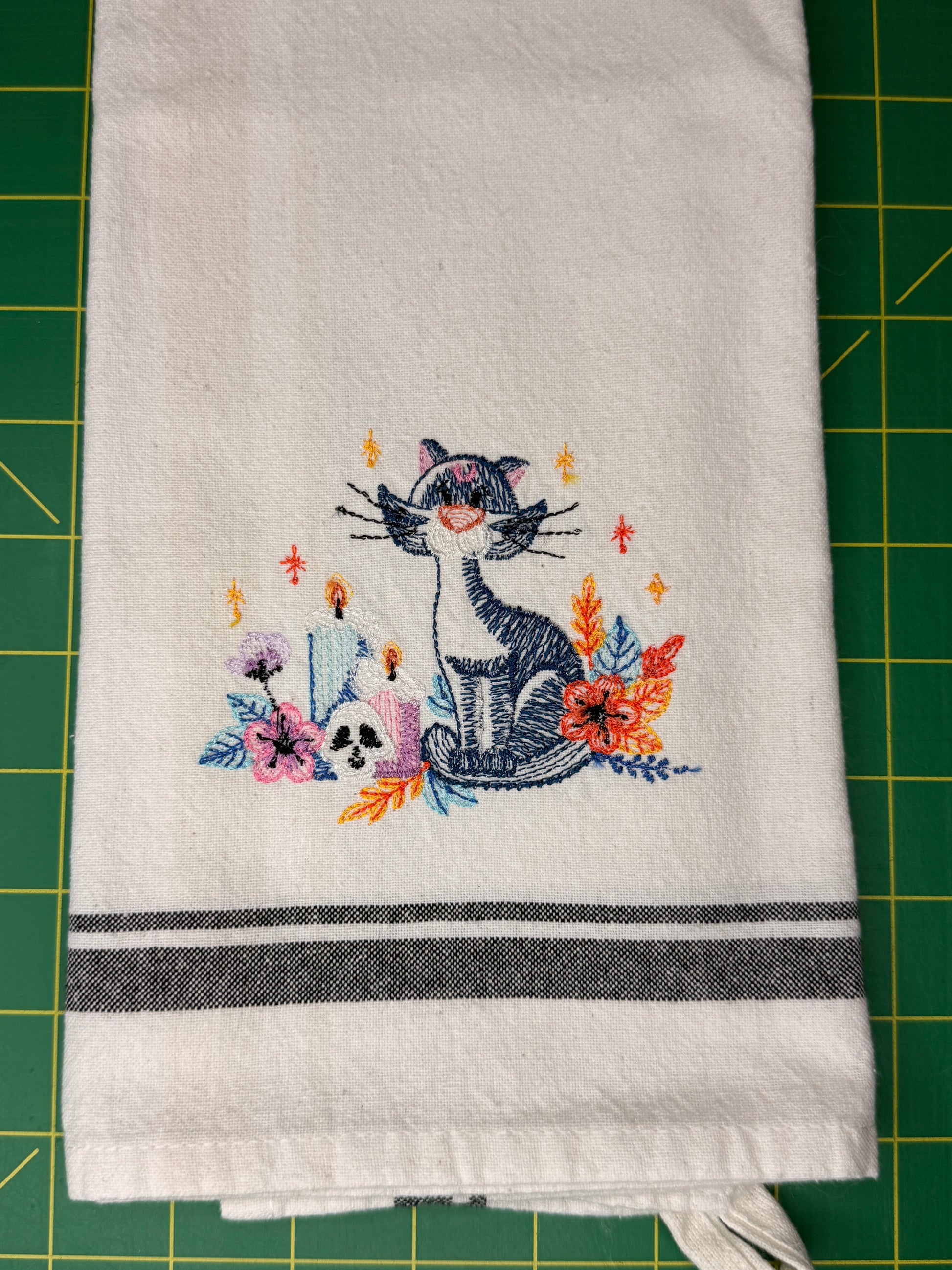 A white kitchen towel with a colorful embroidered design of a cat, flowers, and candles, featuring two dark gray horizontal stripes near the bottom, laid flat on a green cutting mat.