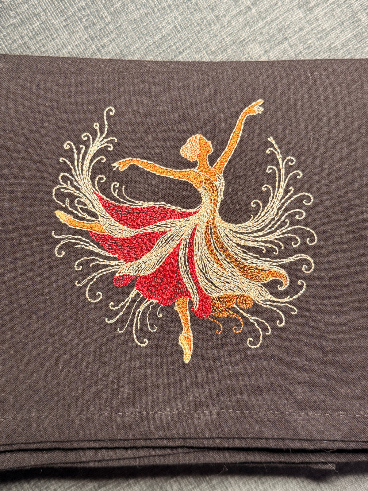 A folded dark black fabric towel featuring an embroidered design of a dancing woman in a flowing red and gold dress with white decorative swirls.