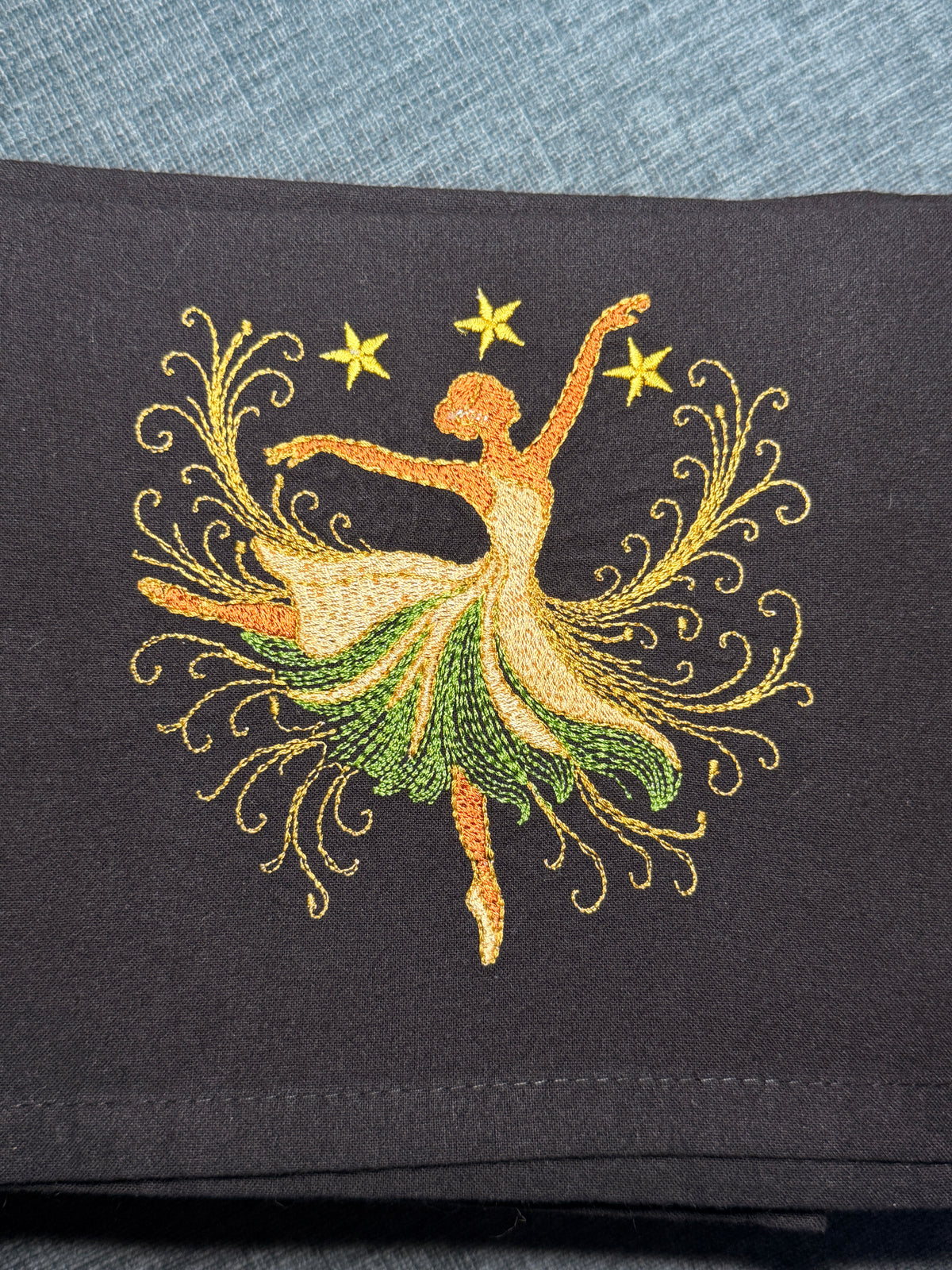 A close-up of a black fabric towel, featuring an embroidered design of a dancing fairy with a green and gold skirt and yellow stars.