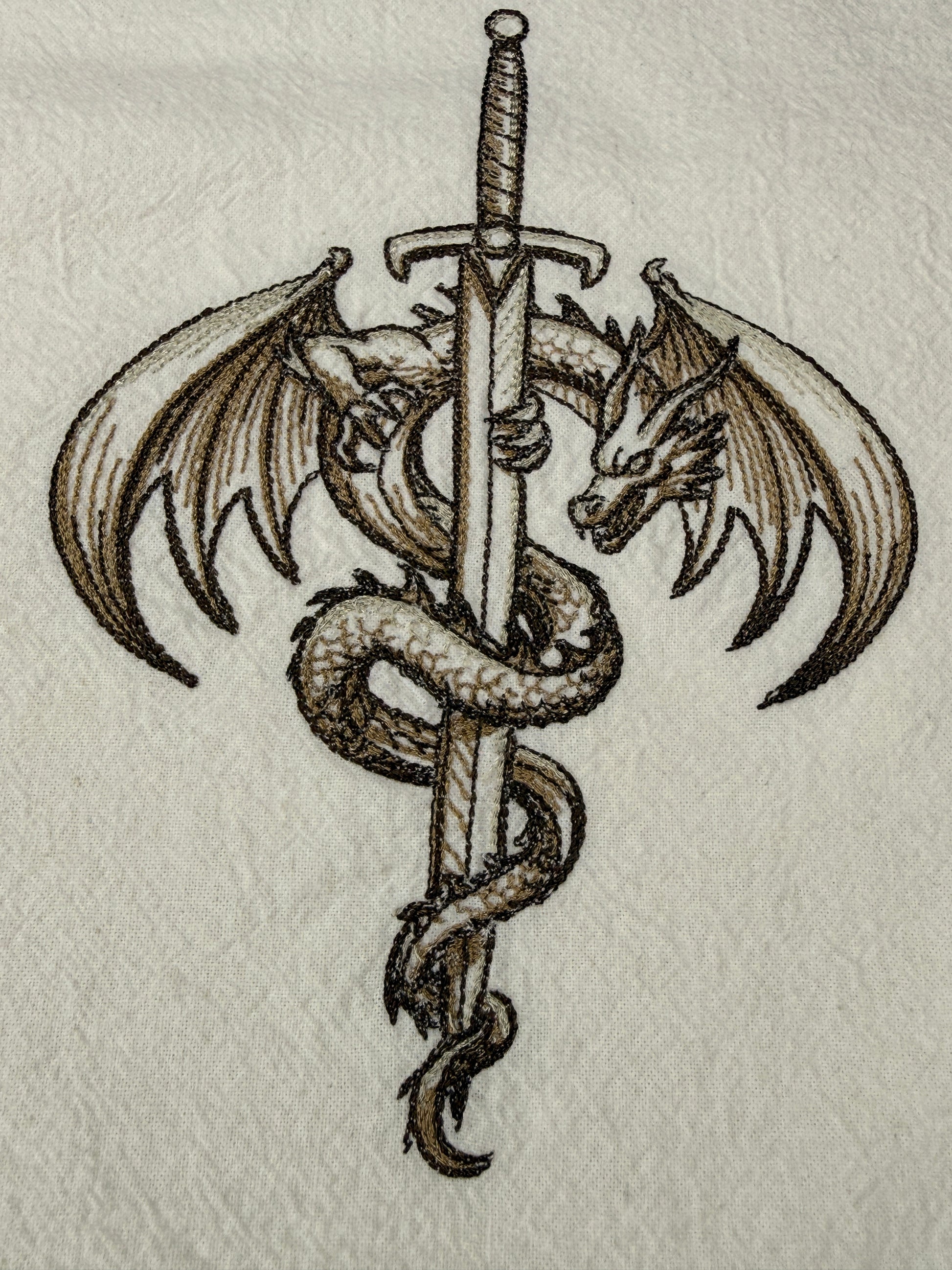A detailed embroidery design of a winged dragon wrapped around a vertical sword, shown on a textured off-white fabric background.