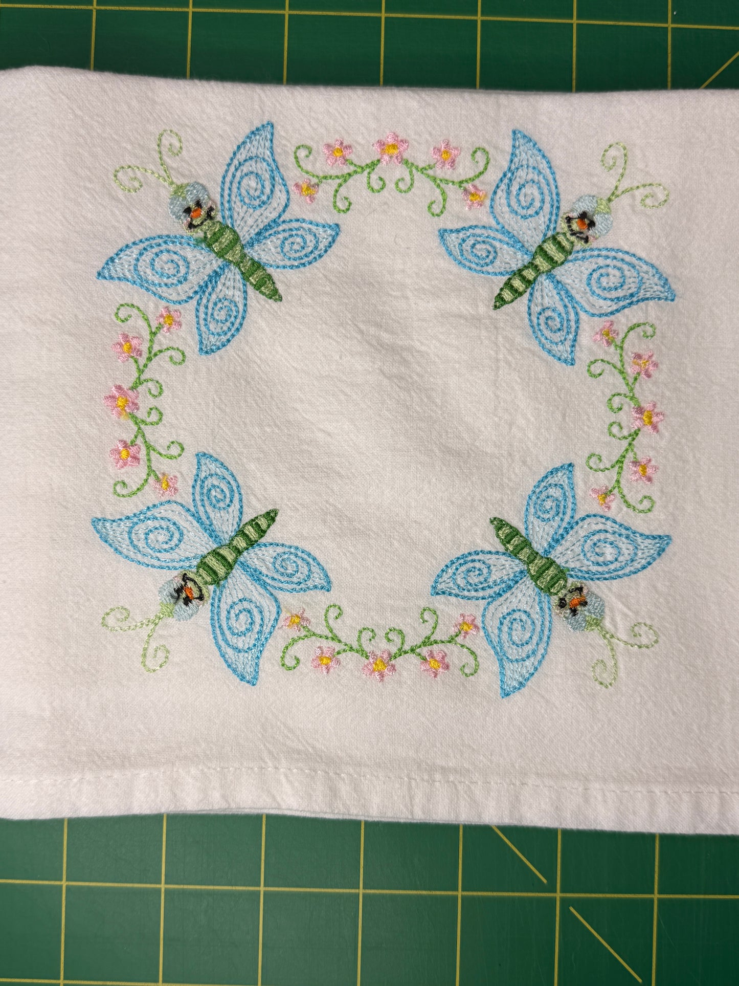 A square white fabric towel featuring embroidered blue butterflies and green floral vines, laid flat on a green cutting mat.