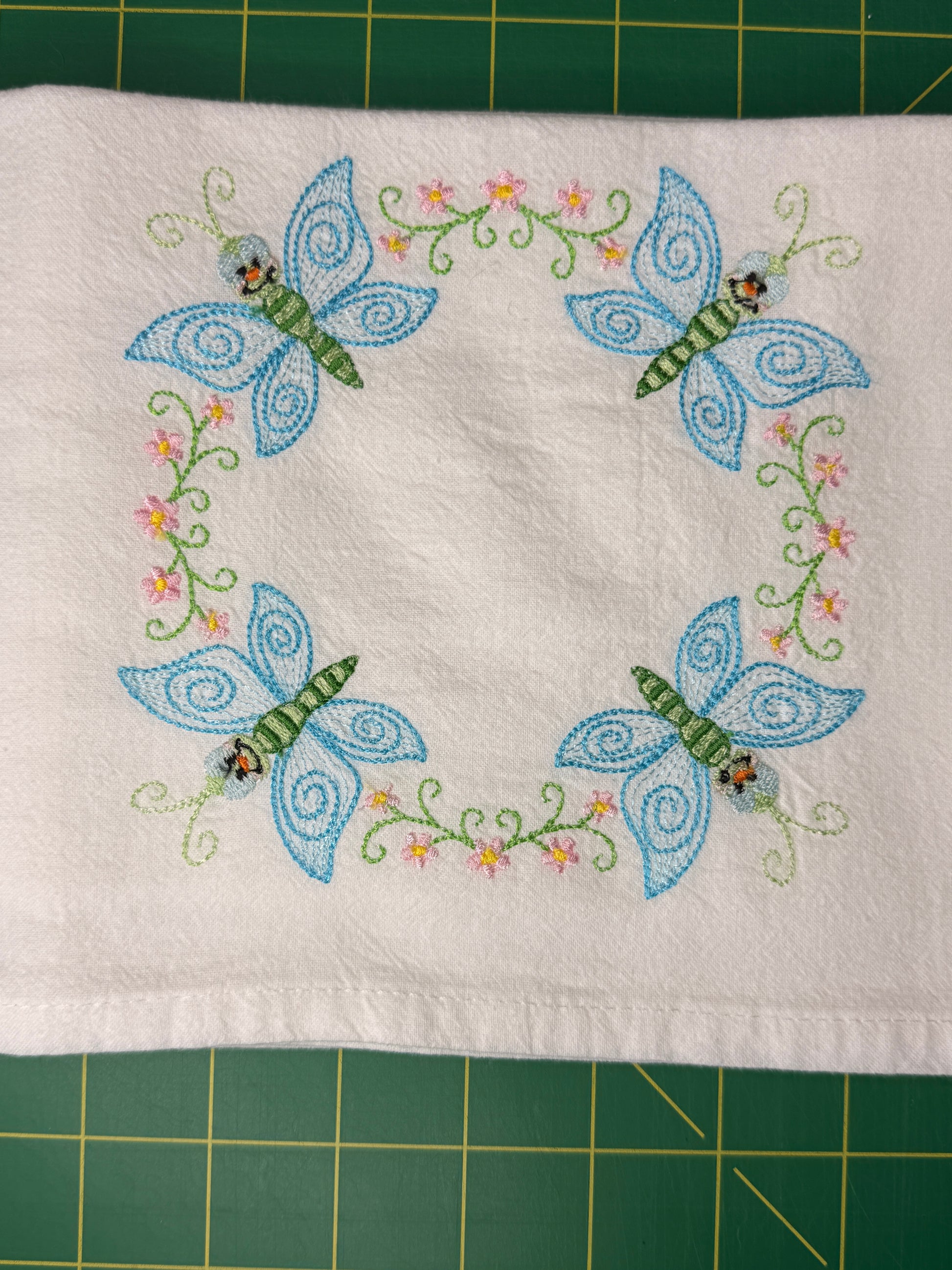 A square white fabric towel featuring embroidered blue butterflies and green floral vines, laid flat on a green cutting mat.