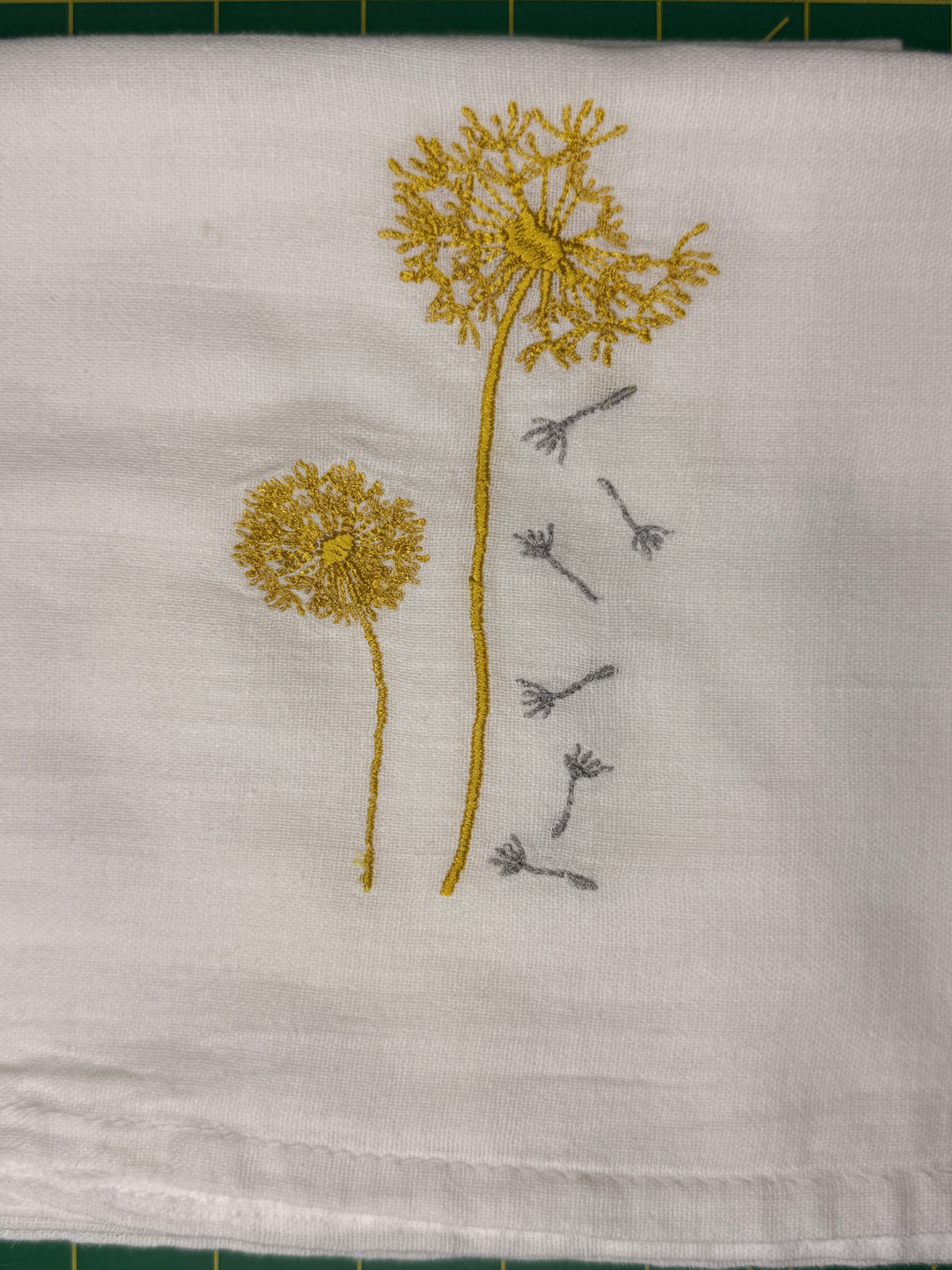 A close-up of a white fabric towel featuring embroidered dandelion designs in yellow and gray.