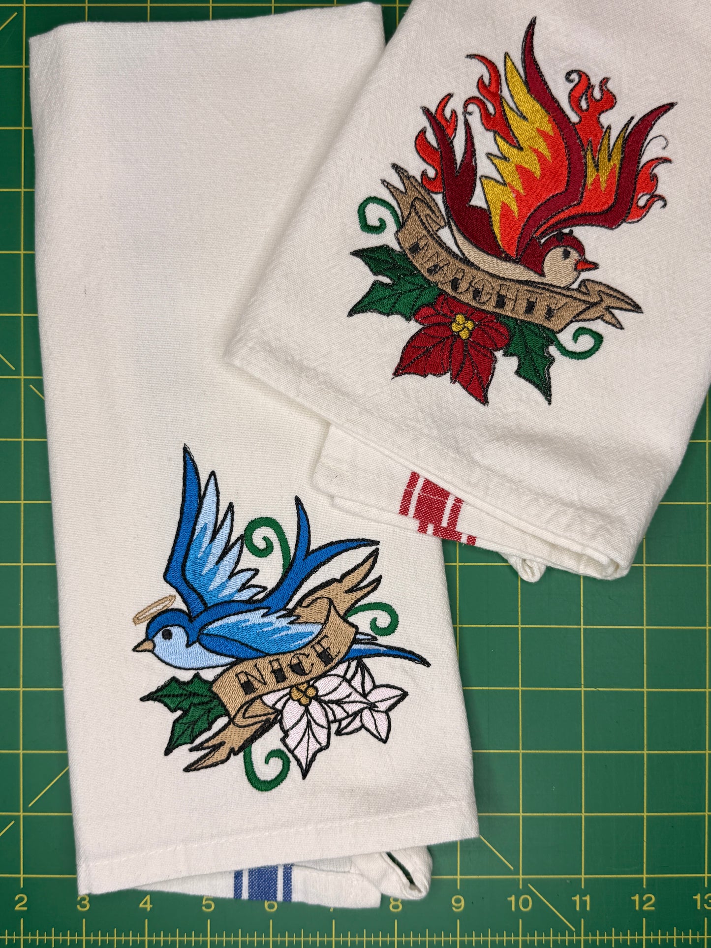 Two white towels with colorful embroidered designs, one featuring a blue bird and the other a red bird with flames, laid flat on a green cutting mat.