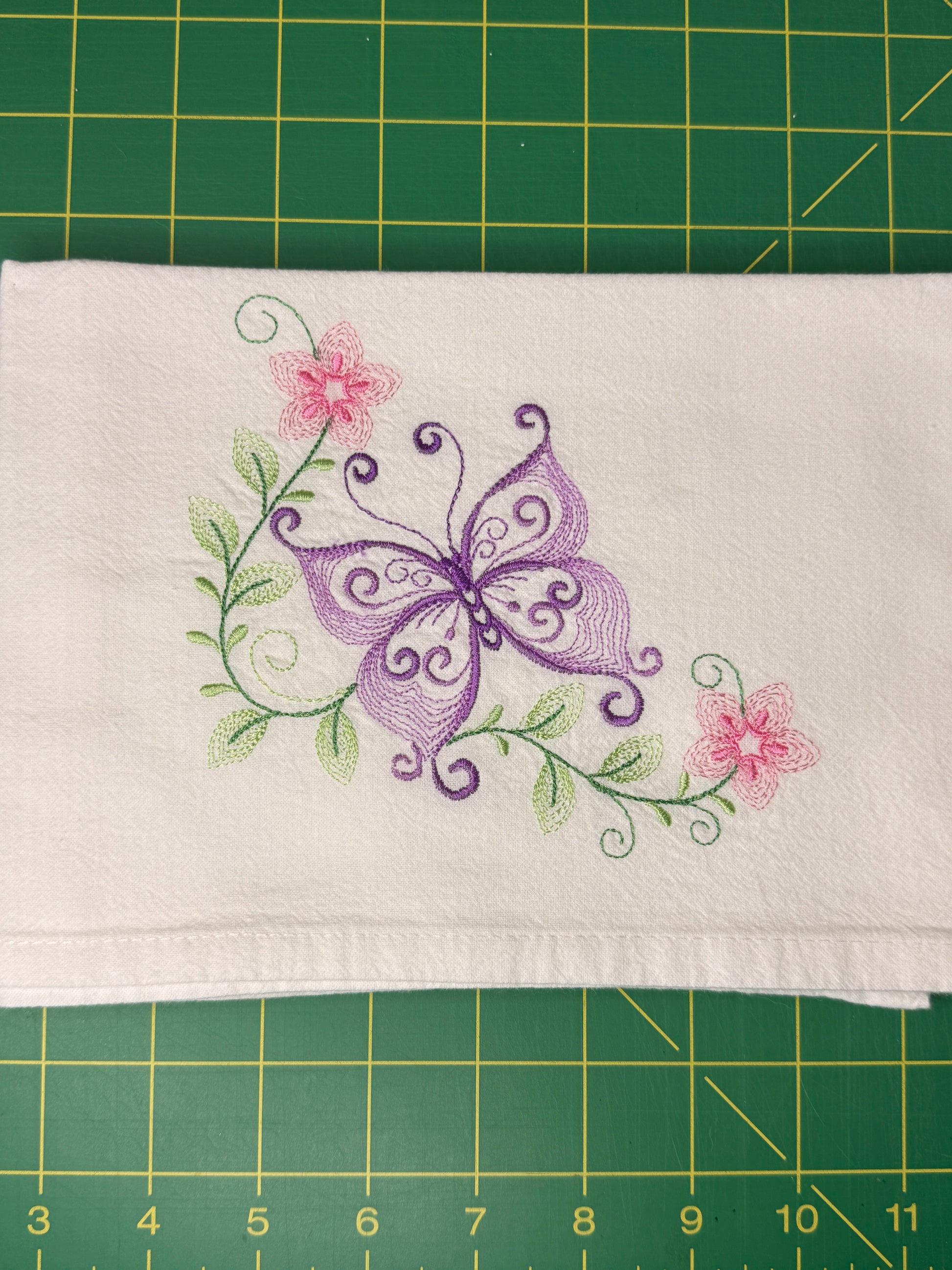 A folded white fabric towel featuring an embroidered design of a purple butterfly, green leaves, and pink flowers, laid flat on a green cutting mat.