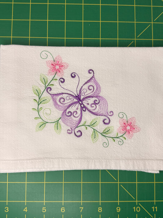A folded white fabric towel featuring an embroidered design of a purple butterfly, green leaves, and pink flowers, laid flat on a green cutting mat.