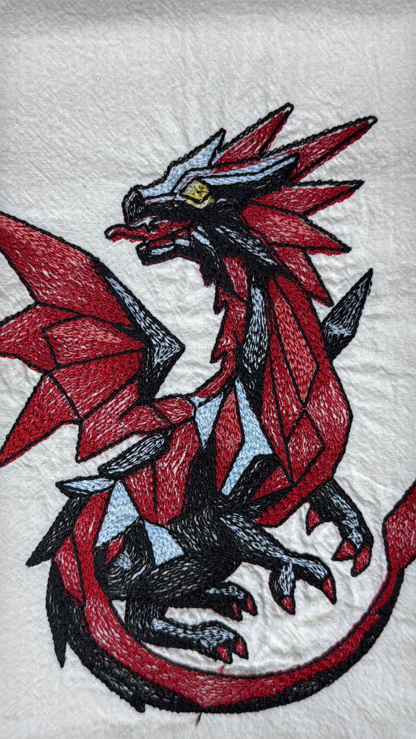 A close-up of a white embroidered fabric featuring a stylized red, black, and gray dragon with small light blue accents and a yellow eye.
