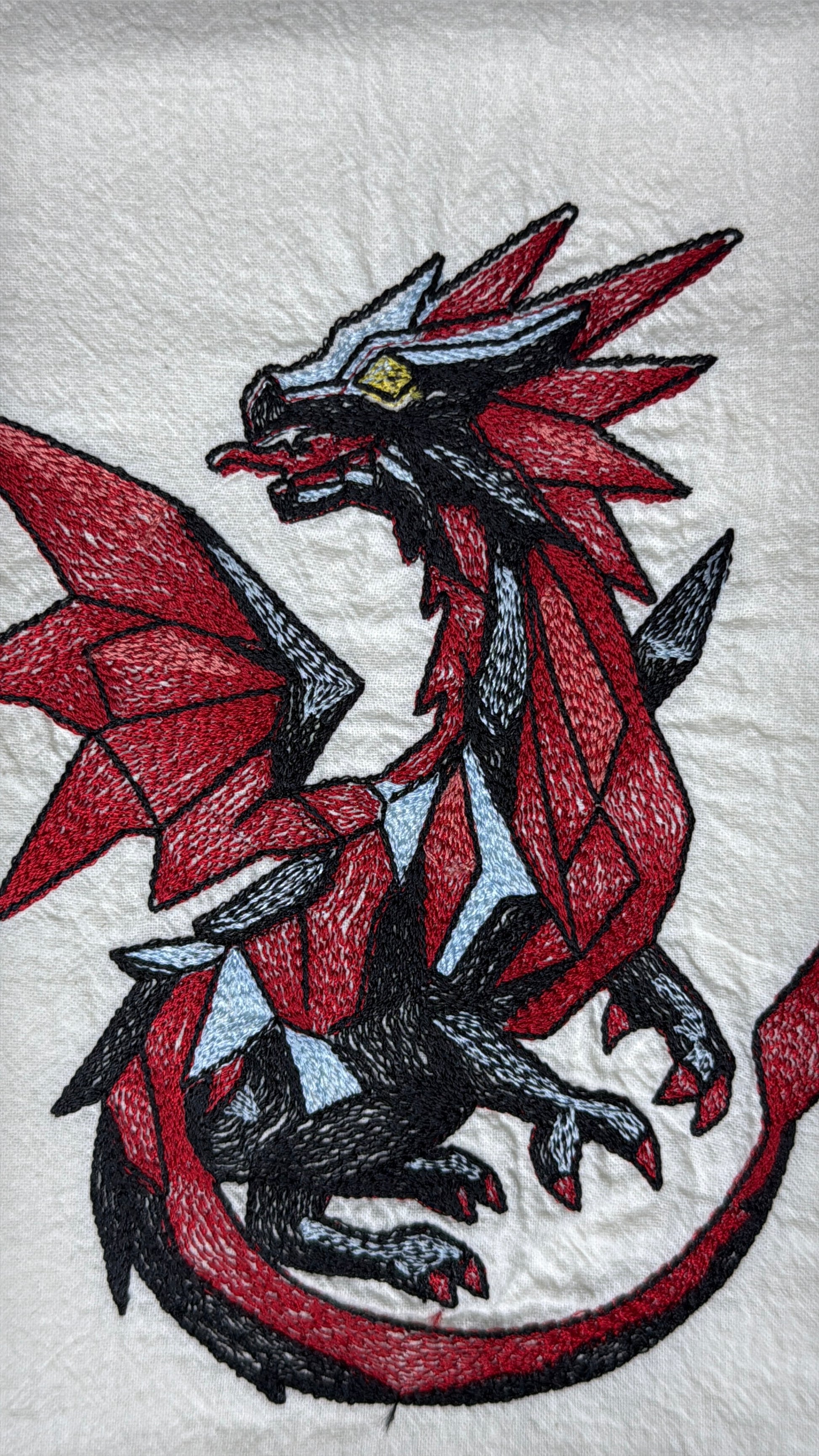 A close-up of a white embroidered fabric featuring a stylized red, black, and gray dragon with small light blue accents and a yellow eye.