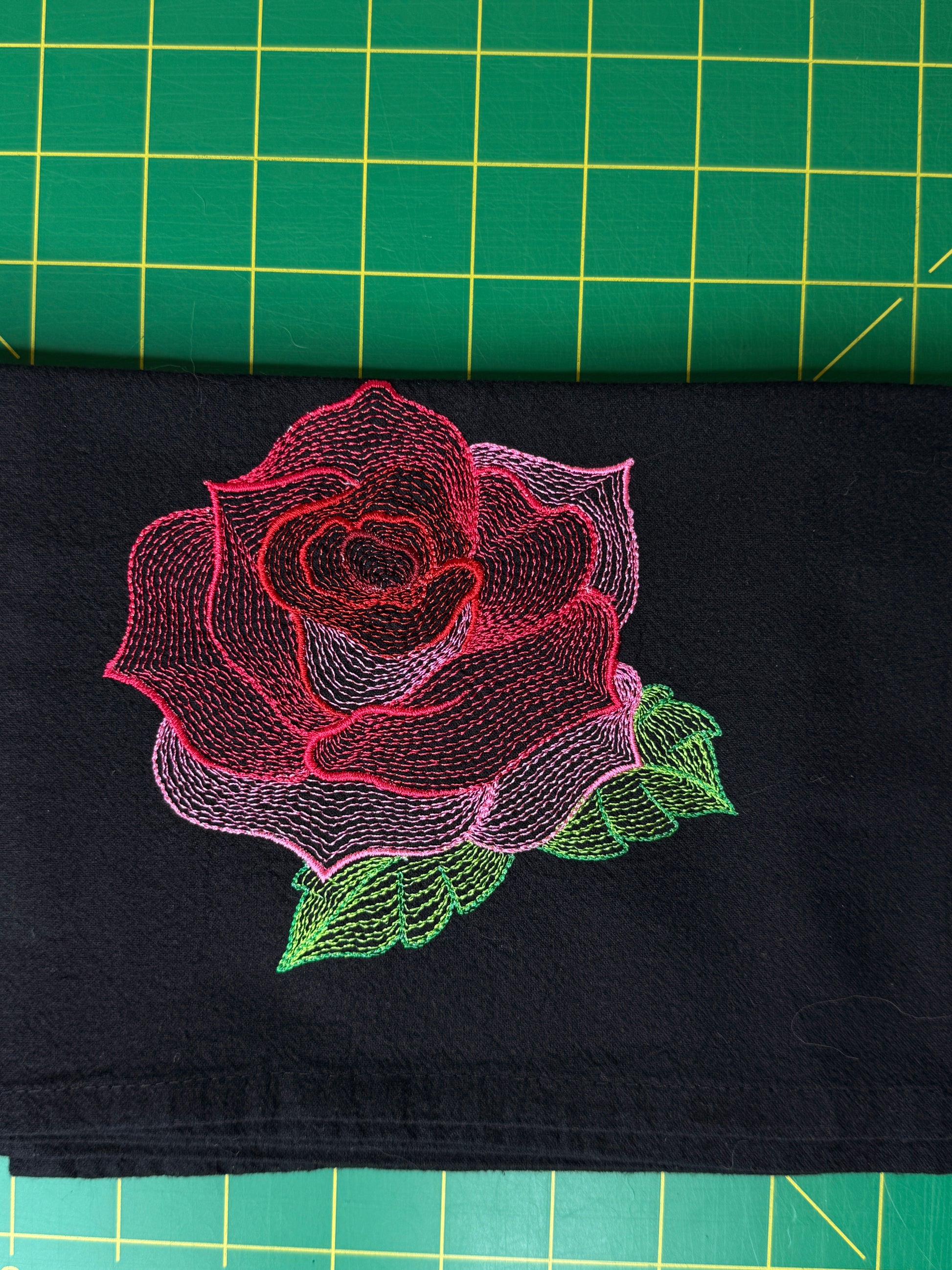 A close-up of a black fabric towel featuring a large embroidered rose design in pink, red, and green.