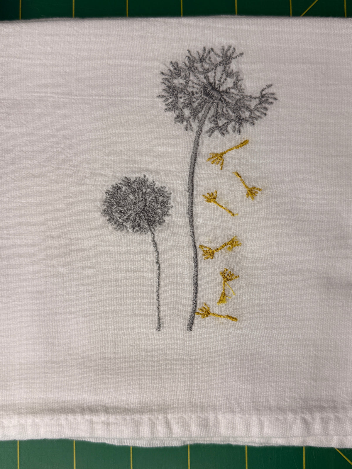 A close-up of a white gauzy fabric towel featuring embroidered dandelions in gray and yellow. The fabric is laid flat on a green cutting mat.