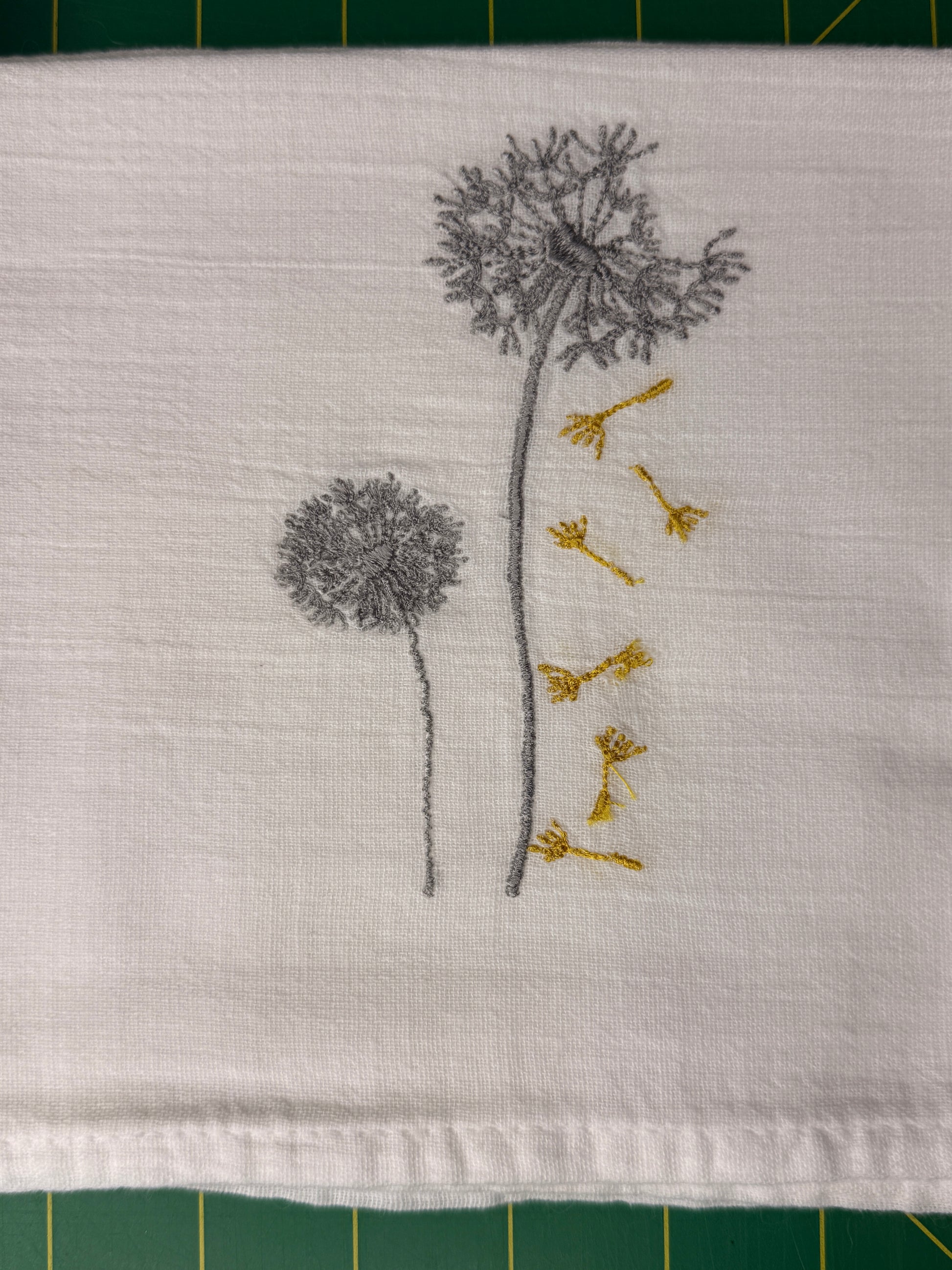 A close-up of a white gauzy fabric towel featuring embroidered dandelions in gray and yellow. The fabric is laid flat on a green cutting mat.