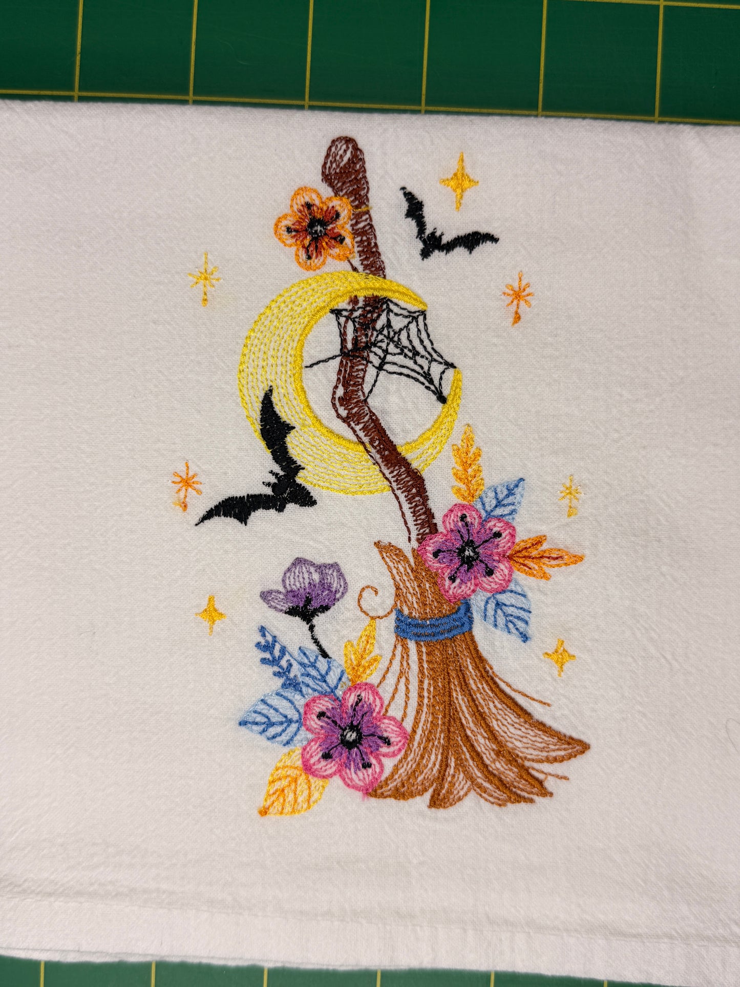 A close-up of a white fabric towel featuring a colorful embroidered witch design with a broomstick, crescent moon, bats, flowers, and stars. The fabric is laid flat on a green cutting mat.