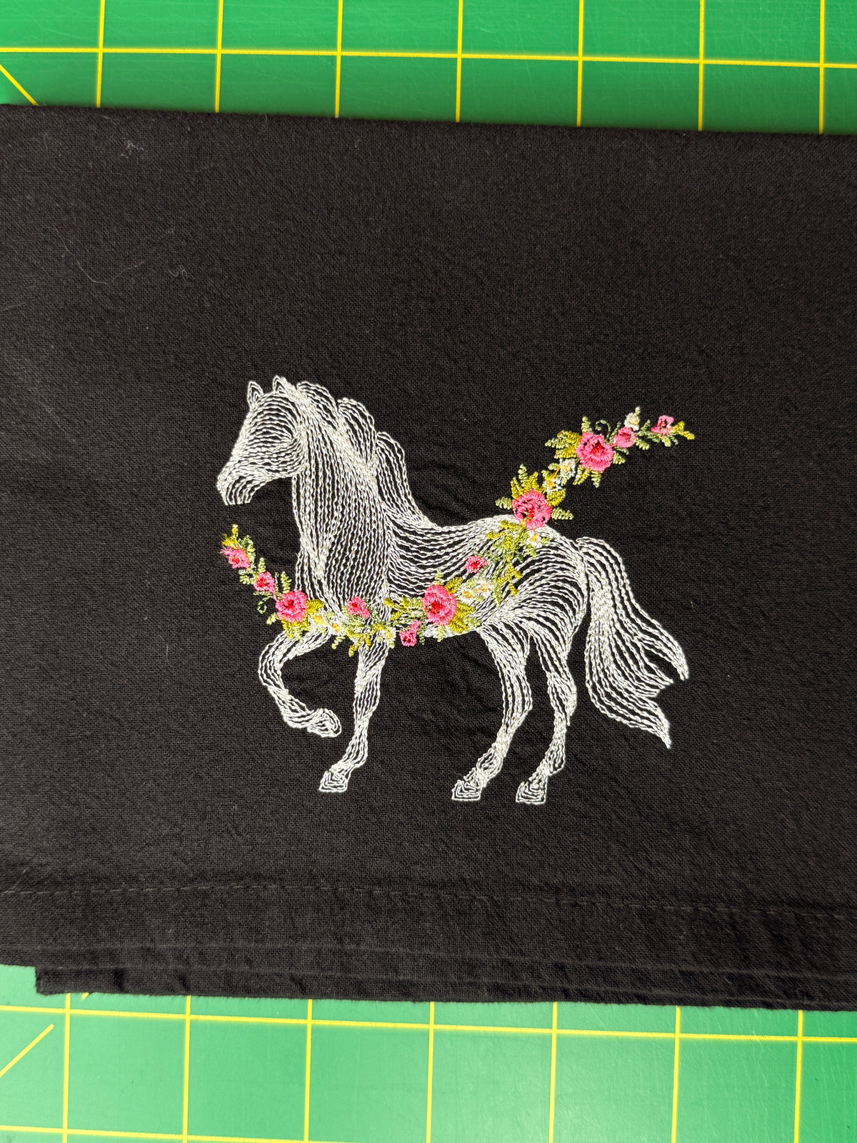 A black fabric towel featuring a white embroidered line-art horse adorned with pink and green floral garlands. 
