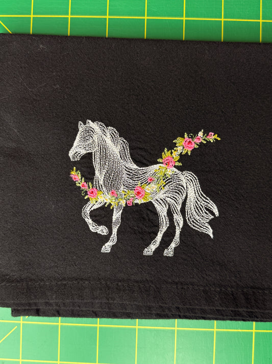 A black fabric towel featuring a white embroidered line-art horse adorned with pink and green floral garlands. 